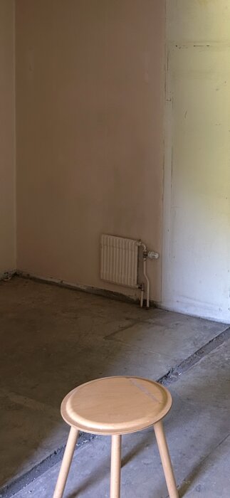 Tom rum med betonggolv och en spårning där en vägg stod, pall i förgrunden och radiator på väggen.