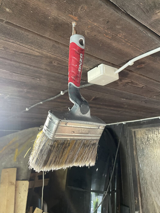 Målarpensel med rött och svart handtag hänger från träbjälke i garaget. Penseln är använd och hänger för att torka eller förvaras mellan målningsarbeten.