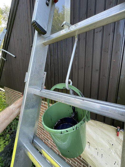 Grön hink med brun färg hänger på stege vid brunmålad husfasad; ett verktyg syns inuti hinken.