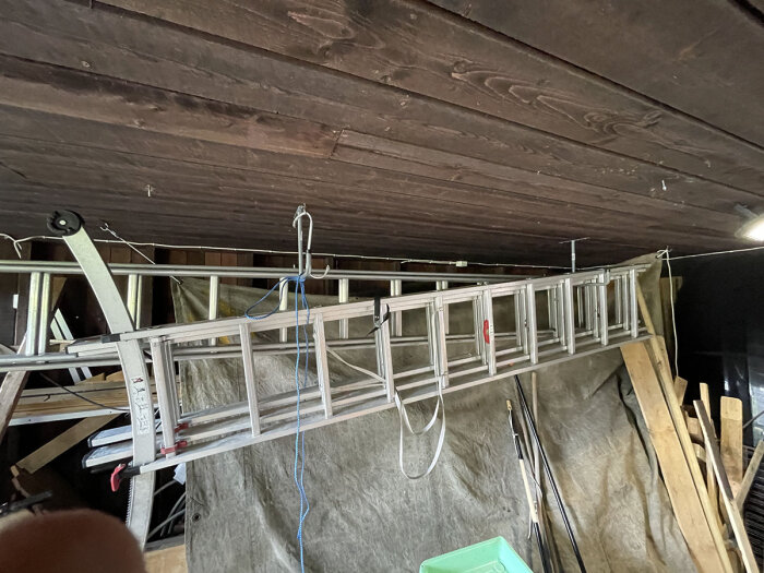 Aluminiumstege förvarad upphängd i ett förråd, omgiven av träbrädor och olika verktyg.