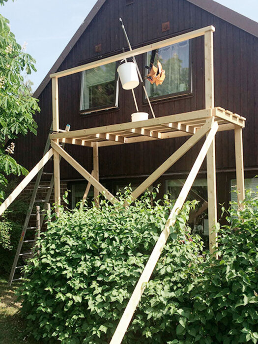 En enkel träställning framför en mörkbrun husfasad, med en stege och en hink upphängd. Ställningen står över en grön buske.