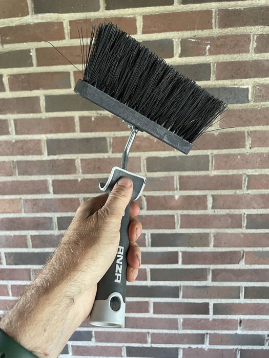 Person som håller en Anza fasadtvättborste mot en tegelvägg, använd för att rengöra fasaden innan målning.