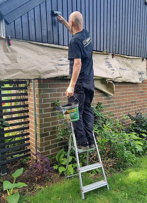 En person målar en hög väggpanel i mörk färg med en roller, stående på en stege, med en plasthink från Cuprinol i handen.