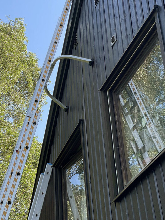 Stege med avståndshållare lutad mot nymålad brunsvart fasad. Klarblå himmel och träd i bakgrunden.