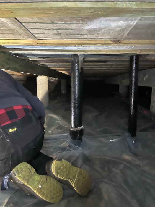 Person inspekterar under hus med avloppsrör i centrum.