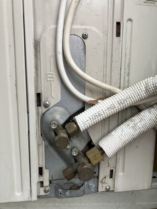 Luft/luft kompressor med ventiler och muttrar på en vit enhet. Två rör anslutna, ett med vit isolering.