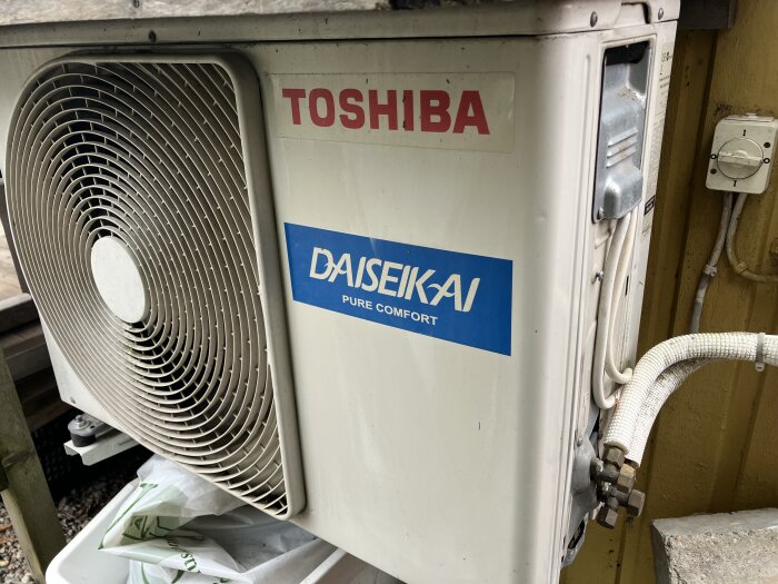 Luft/luft-kompressor med etiketter "Toshiba" och "Daiseikai Pure Comfort", två ventiler längst ner, monterad på väggen utomhus.