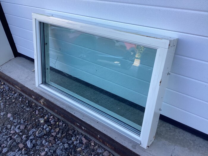 Fast fönster med sprucken innerglasruta och vit träkarm, placerat utomhus mot en vägg av vit panel.