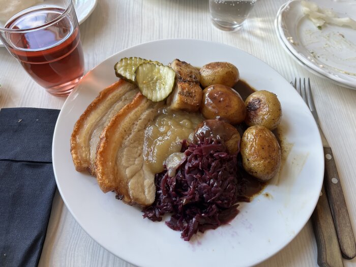 Tallrik med flæskesteg, potatis, rödkål, skysås och inlagd gurka. Glas med röd dryck bredvid tallriken.
