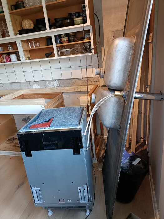 Diskbänk i metall lutar mot vägg, diskmaskin står framför i renoverat kök med halvklädd vägg och öppna köksskåp fyllda med porslin.