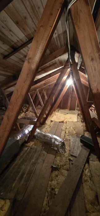 Vind med träbjälkar, isoleringsmaterial på golvet och en synlig ventilationskanal i metallfolie.