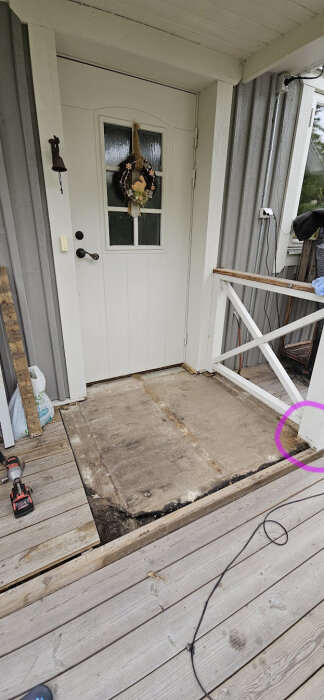 Framsidan av en husentré med vit dörr, farstukvist med träplankor och skadad betongkant markerad i ett hörn. Verktyg och material syns på marken.