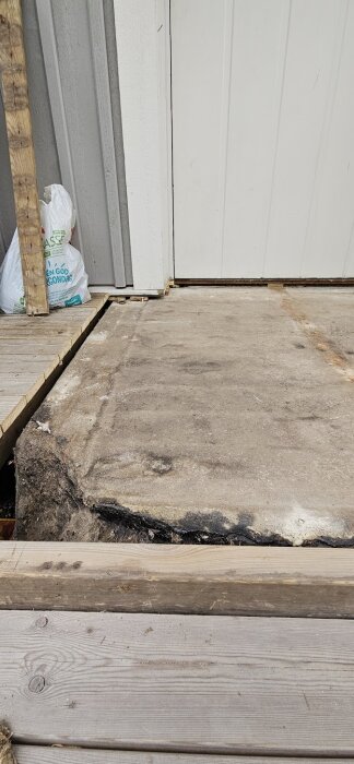 Sprucken betongyta vid entré med träbrädor omkring och vit dörr i bakgrunden. Synlig skada indikerar vatteninträngning och behov av reparation.