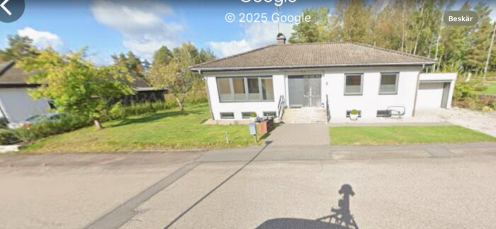 Framsida av ett hus med gräsmatta och uppfart, där en stenmur planeras att byggas. Skugga av en kamera syns på vägen.