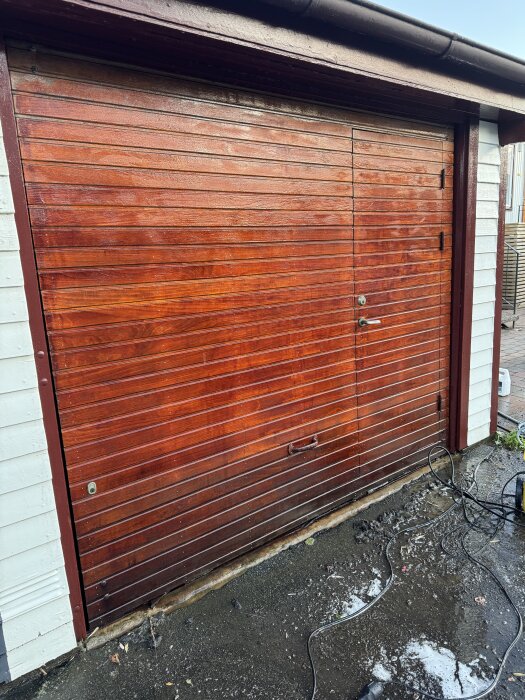 Rengjord garageport i trä från 1962 med rödbrun finish, omgiven av vitmålade väggar och en mörk ram, kablar synliga på marken.