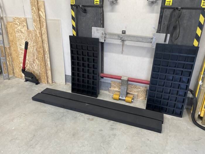 Byggmaterial och verktyg mot en vägg, inklusive svart gummimattor, plywoodskivor och en stång med gula hjul på ett betonggolv.