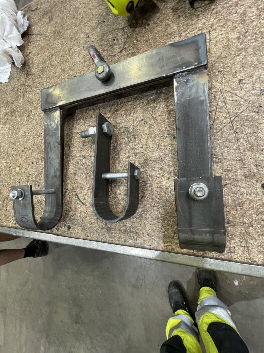 Metallfäste med tre krokar på arbetsbänk, verktyg synligt i bakgrunden.