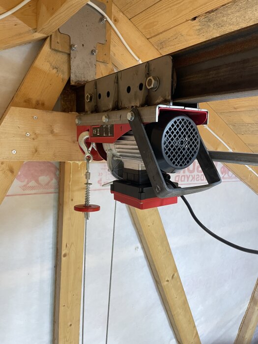 Elektrisk vinsch monterad på en träbalk i ett garage eller arbetsutrymme. Synliga kablar och stålvajer med röd spärrknapp hänger ner.
