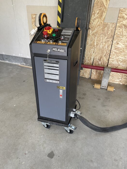 Verktygslåda från PELA Tools på hjul med kontrollpanel, strömkabel och märkning för 230V, placerad i ett rum med verktyg och skivmaterial.