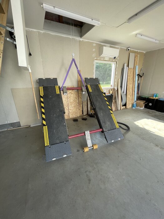 Lyftplattform i en garageverkstad, två ramper med gul-svarta markeringar, upphängd på en lila rem, omgiven av byggmaterial och verktyg.