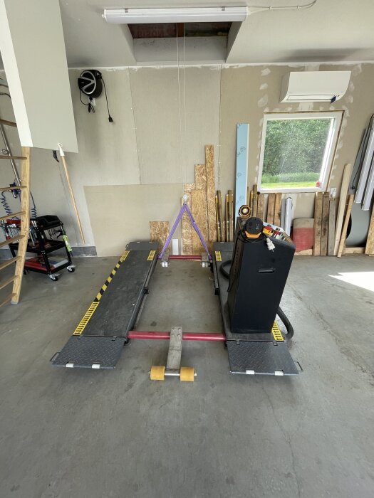 Lyftplattform i ett garage med verktyg och förvaringsutrustning i bakgrunden och ett fönster som visar grönska utanför.
