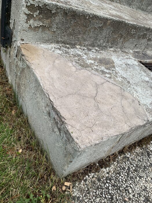 Gamla lagningar och sprickor i en betongtrappa nära marknivå, med flagnande yta och omgiven av gräs och grus.