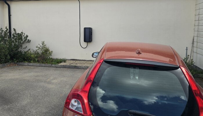 Röd bil parkerad framför en vit vägg med en laddbox monterad, svart rördragning synlig; växtlighet vid sidan av bilen.