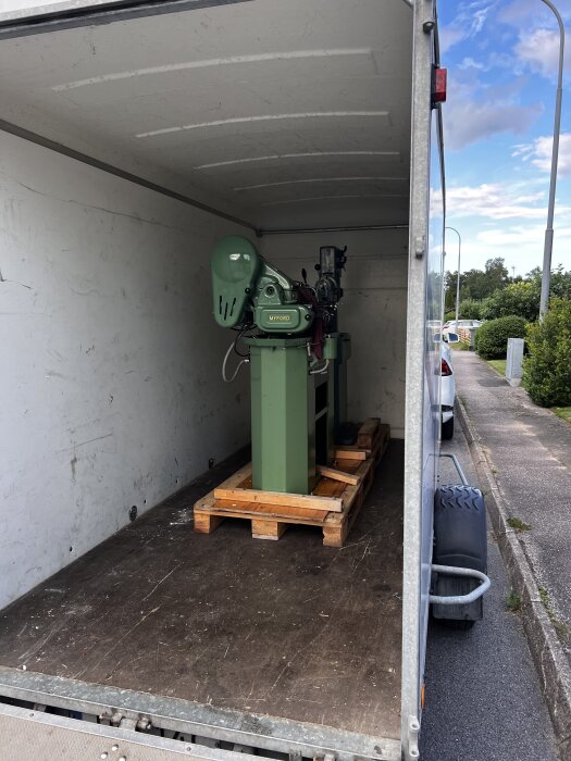Grön Myford-maskin på en träpall i en lastbilstransport, parkerad vid en trottoar under blå himmel.