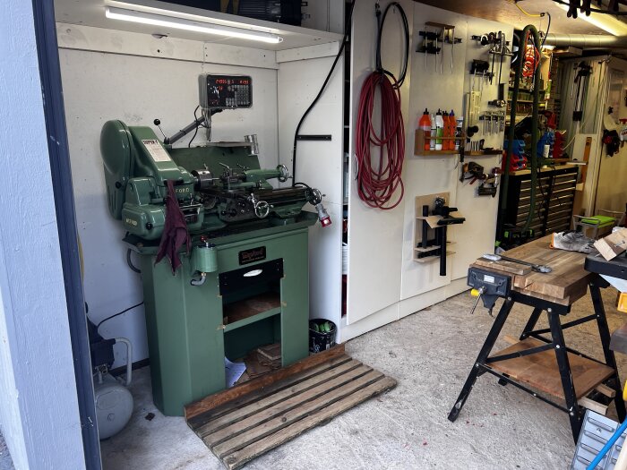 Grön svarvmaskin i ett verkstadsutrymme, omgiven av verktyg på väggar och en arbetsbänk.