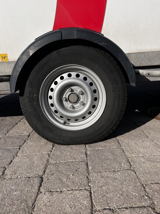 Närbild på ett däck och hjul av ett fordon, parkerat på marksten, med en del av ett rött och vitt fordon synligt i bakgrunden.
