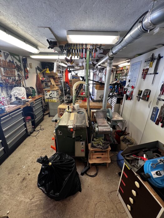 Verkstad fylld med verktyg, bänkar, skåp och utrustning; avfallspåse på golvet och välfyllda hyllor i bakgrunden. Taket har belysning och ventilation.