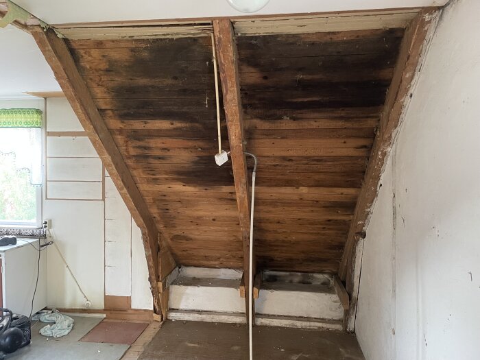 Interiörbild av övervåning i äldre hus med synligt träbjälklag i taket och delar av väggen. Målade och omålade ytor, elledning synlig.