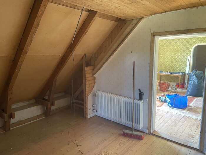 Övervåning i äldre hus under renovering med synliga takbjälkar, radiator och städutrustning. Dörröppning leder till ett rum med byggmaterial.
