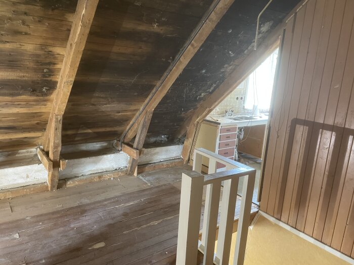 Gammalt vindsutrymme med träbjälkar och synligt tak, delvis renoverat. En spis och handfat i bakgrunden, förberett för isolering.