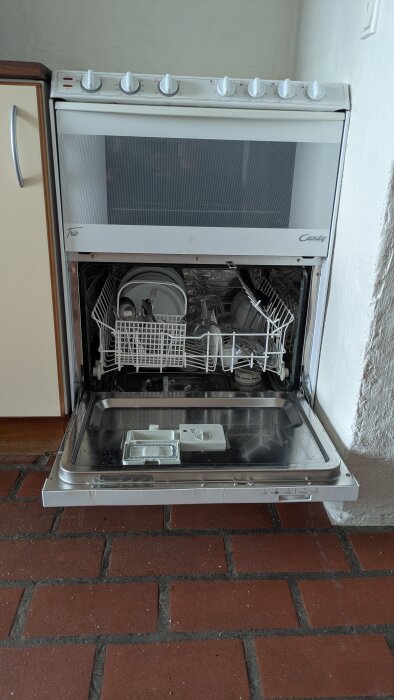 Diskmaskin med öppen lucka, fylld med tallrikar och bestick, installerad intill köksskåp på klinkergolv.