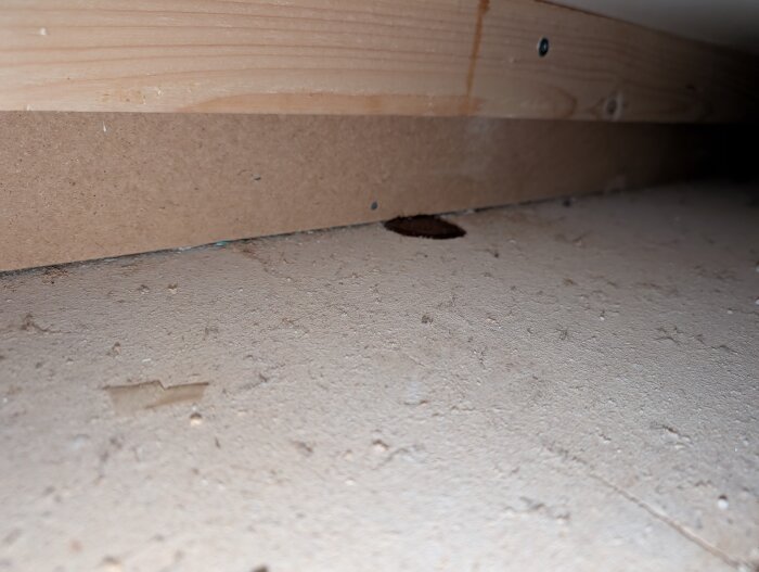 Mörk fläck på golvspån under ett köksskåp i ett trähus från 1983. Fläcken liknar sirap och finns vid en träplankskant. Inga synliga spår ovanför.