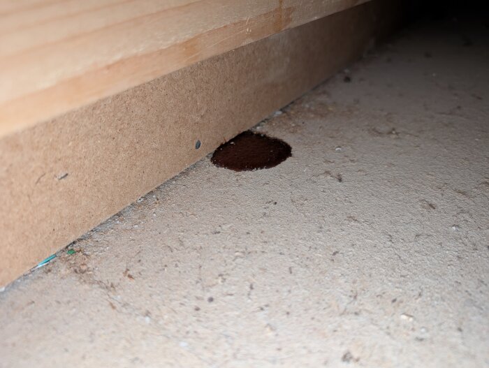 Mörkfläck på golvspån, liknar halvtorkad sirap, under köksskåp i hus från 1983. Fläcken har droppform och ser ut att ha runnit ovanifrån.