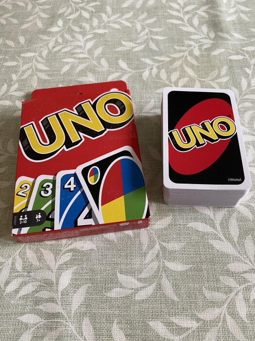 Kortleken UNO med en öppen låda och en hög med kort visas på ett bord med bladmönstrad duk.