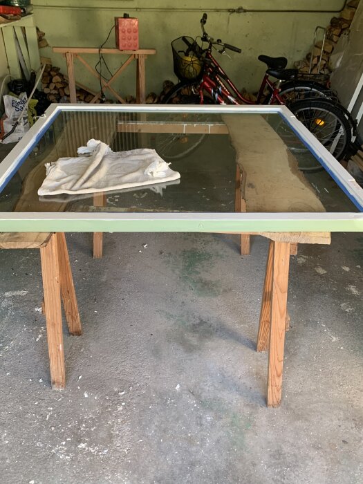 Fönsterram med glas placerad på två bockar i ett garage, med en trasa ovanpå och cyklar samt ved i bakgrunden.