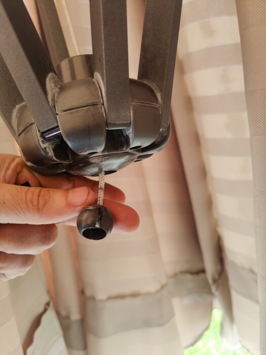 Hand som håller i ett snöre med en kula i slutet, del av en persienne eller gardinmekanism i bakgrunden.