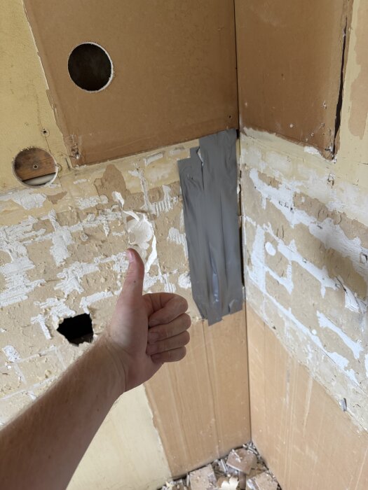 En hand ger tummen upp framför en vägg där kakel och delar av gipsskiva tagits bort. Väggytan är delvis tejpad med silvertejp.