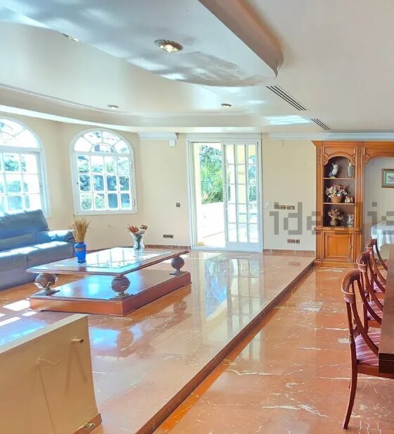 Vardagsrum med marmorgolv och höjdskillnad; soffa, soffbord och vitrinskåp synliga, stora fönster och altandörr i bakgrunden.