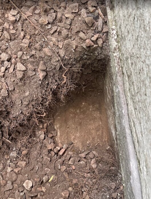 Grävd grop runt en betongplatta, exponerad jord och stenar, vid en tillbyggnad från 80-talet med bekymmer om fukt underifrån.