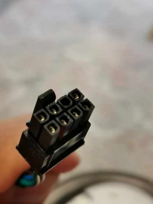 8-pin elkontakt i närbild för anslutning av fläkt, hålls i en hand. Bakgrunden är suddig med ljusa färger.