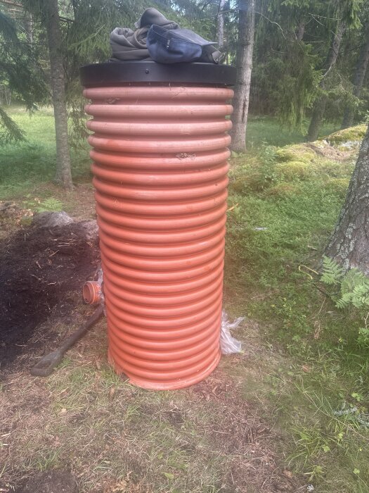 Orange biofiltertunna i skogsmiljö, topplock av och kläder ovanpå, redo för installation i sommarstugan.
