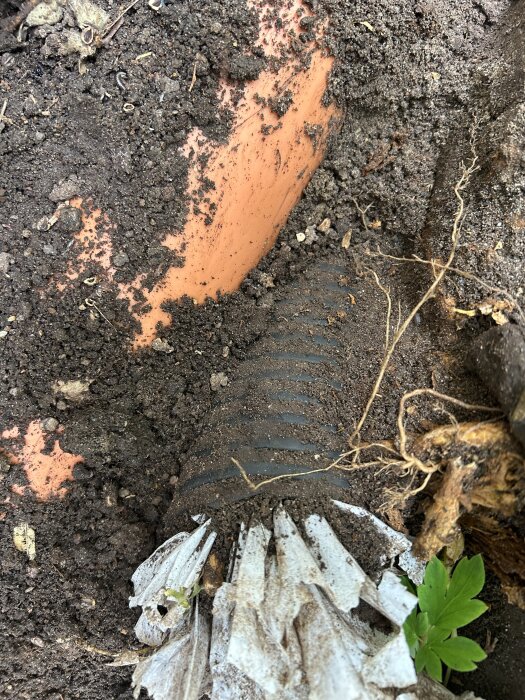 Svart igentäppt rör i jord bredvid orange rör, samt ogräs och växtrester. Rören är inte kopplade; okänd funktion.
