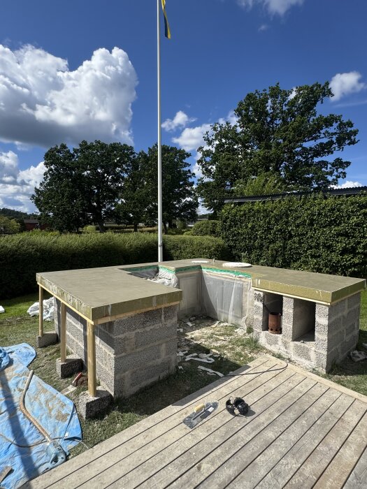 Betongskiva nygjuten utomhus med stödkonstruktion i trä och stenblock, omgiven av natur och flaggstång under blå himmel.