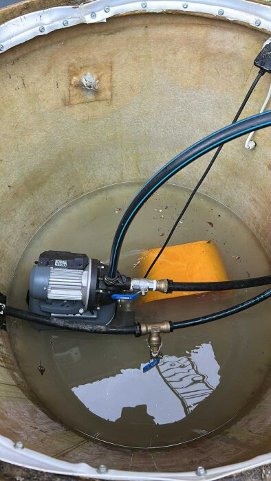 Pump i glasfiberbrunn med vatten intill, orange flytboj bredvid, slangen ansluten.
