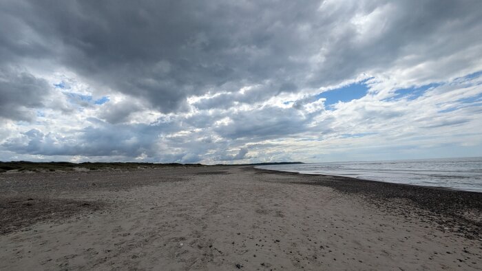 Sandstrand med mörka skyar och havet till höger, molnigt väder.