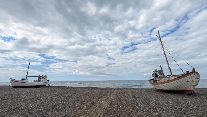 Två nyfiskebåtar står på en stenig strand med havet och molnig himmel i bakgrunden.